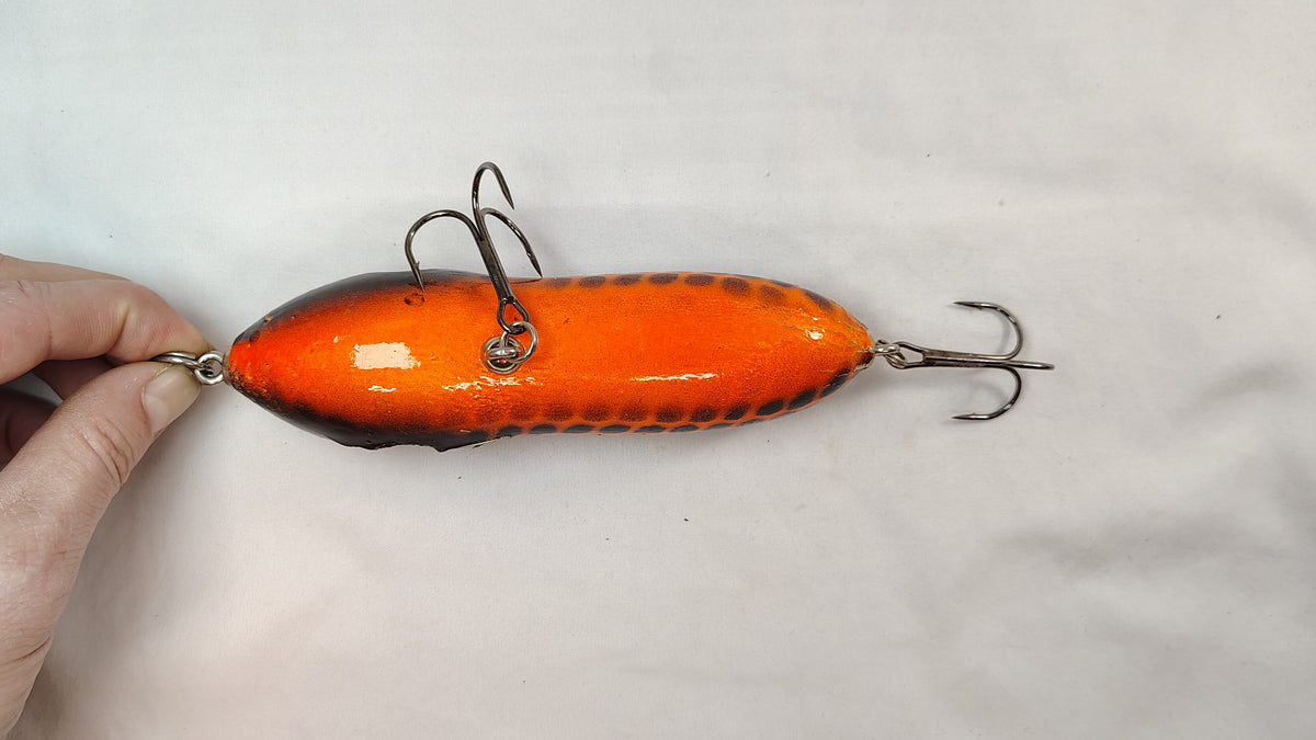 Orange fishing lure with metal hooks held by a hand on a light gray background
